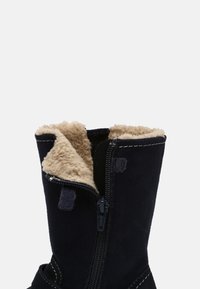 Botte d'hiver en daim noir avec doublure en polaire beige, partiellement dézippée pour montrer l'intérieur doux et les détails de couture blanche.