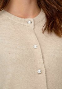 Beige Strickjacke aus weichem Strickstoff, mit einem runden Ausschnitt und drei perlenartigen Knöpfen an der Vorderseite. Glatte, strukturierte Oberfläche.