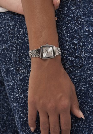 Handgelenk mit einer silbernen, quadratischen Armbanduhr und einem verlinkten Metallband, das auf einem strukturierten blauen Stoffhintergrund liegt.