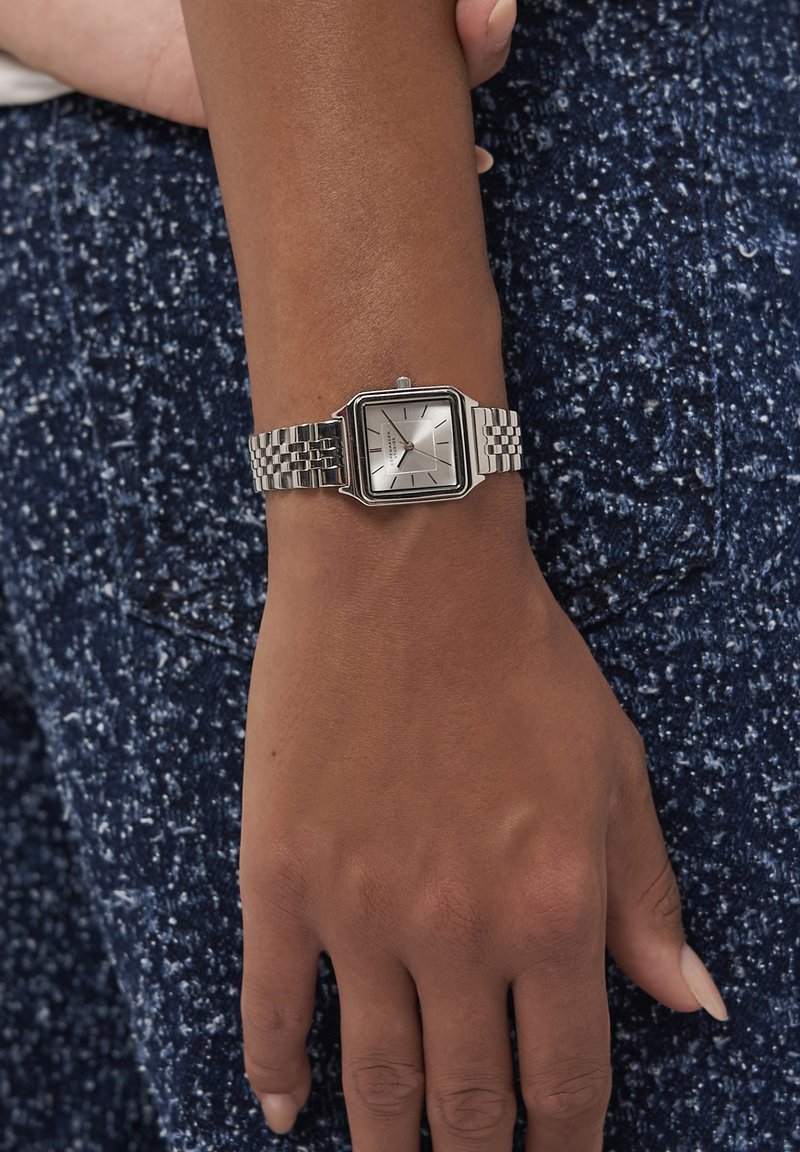 Handled med en silver fyrkantig klocka och länkad metallband som vilar på en strukturerad blå tygbakgrund.