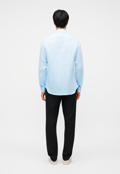 Άνδρας που στέκεται πλάτη, φοράει ανοιχτό μπλε πουκάμισο με μακριά μανίκια, μαύρο παντελόνι και λευκά αθλητικά παπούτσια, μπροστά σε απλό φόντο.