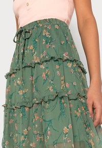 Falda tierna verde con detalles de volantes. La tela presenta flores rosas y amarillas con tallos verdes, cintura ajustable con cordón.