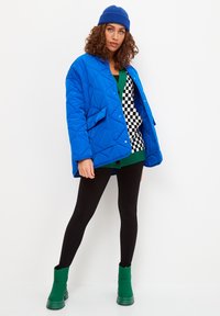 Töltött kék, túlméretezett kabát, alatta zöld-fekete kockás pulóver, fekete leggings és zöld bokacsizma.