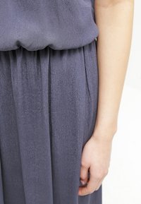 Grå klänning med mjuk textur, har en samlad midja och veckad kjol; hand synlig vid sidan, som visar detaljer av det släta tyget.