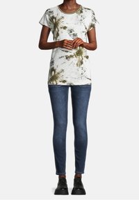 Biała koszulka z krótkim rękawem w zielone wzory tie-dye, okrągły dekolt, zestawiona z dopasowanymi niebieskimi dżinsami i czarnymi butami.
