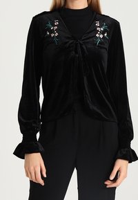 Svart sammetstopp med knytning framtill, broderade blommor på bröstet och utställda ärmar. Mjuk textur med slät finish.