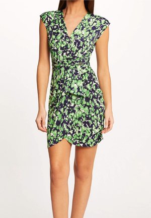 Vrouw draagt een korte mouw, groen-zwart bloemen wikkeljurk met V-hals en asymmetrische zoom, staand tegen een effen achtergrond.