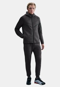 Giacca sportiva grigia con cappuccio e chiusura con zip, abbinata a pantaloni della tuta coordinati. Tessuto liscio, design aderente, cuciture evidenziate e tasche anteriori.