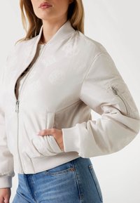 Giacca bomber beige chiaro con una texture liscia, polsini a costine, motivi in rilievo sottili e tasca con zip sulla manica.