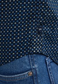 Camicia navy blu con motivo a diamante in azzurro chiaro e crema, caratterizzata da un'etichetta laterale, abbinata a jeans in denim blu e un dettaglio in metallo a forma di bottone rotondo.