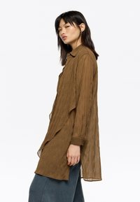 Brown, loosely woven, long-sleeve shirt with a buttoned collar, featuring a layered design and side slits, worn over blue pants.