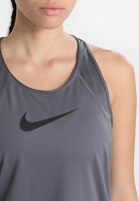 Šedý sportovní tílko vyrobené z lehkého a prodyšného materiálu, které má černé logo Nike na hrudi a kontrastní švy.