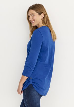 Smilende kvinde med langt lyst brunt hår iført en blå bluse med trekvartærmer og mørkeblå jeans, stående mod en hvid baggrund.