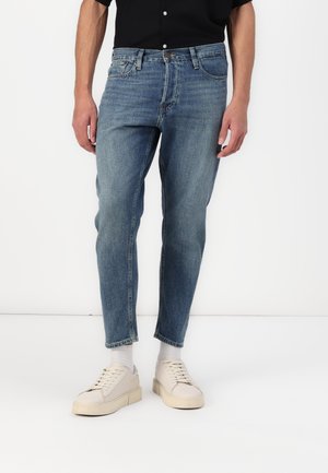 Person trägt mittelblaue Slim-Fit-Jeans, weiße Socken, hellbeige Sneaker und ein schwarzes kurzärmeliges Hemd und steht vor weißem Hintergrund.