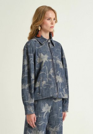 Donna che indossa una giacca con zip a fantasia floreale blu e pantaloni ampi abbinati, accessoriati con orecchini lunghi a nappina arancione.