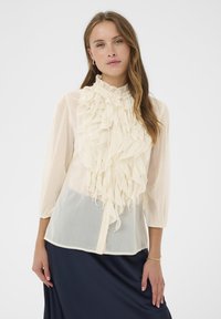 Crème blouse met gerimpeld voorpand, opstaande kraag en knoopsluiting. Driekwart mouwen, doorschijnende textuur en zachte drapering. Gecombineerd met een donkerblauwe rok.
