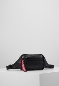 Musta synteettinen vyölaukku, jossa on teksturoitu pinta, varustettuna vetoketjullisella taskulla, säädettävällä hihnalla ja punaisella "POISTA ENNEN LENTOA" -tarralla.