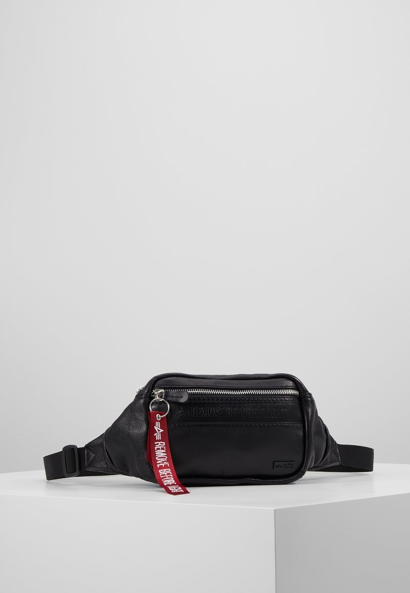 Musta synteettinen vyölaukku, jossa on teksturoitu pinta, varustettuna vetoketjullisella taskulla, säädettävällä hihnalla ja punaisella "POISTA ENNEN LENTOA" -tarralla.