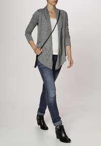 Grå cardigan med öppen front, skiktad design och långa ärmar, bärs över en vit topp och kombineras med blå jeans och svarta ankelstövlar.