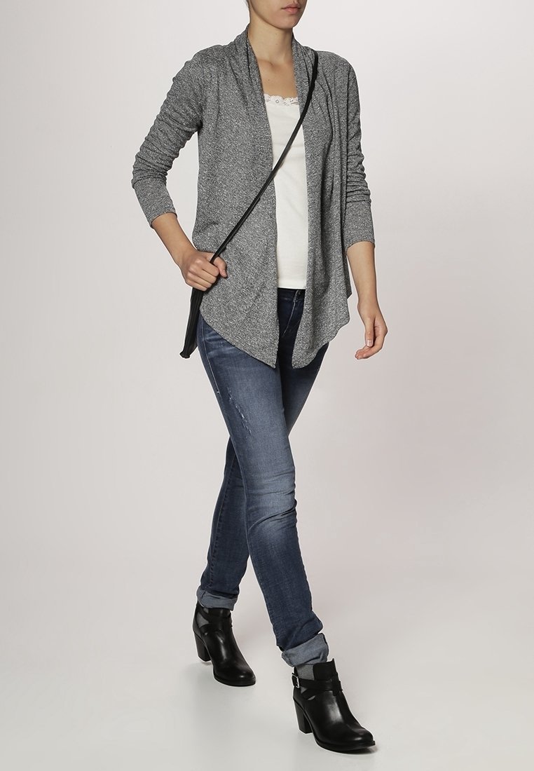 Grå cardigan med öppen front, skiktad design och långa ärmar, bärs över en vit topp och kombineras med blå jeans och svarta ankelstövlar.