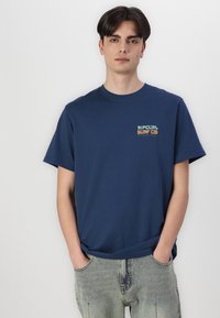 Marineblå bomulds t-shirt med et logo i brystet i grøn og orange, korte ærmer og en afslappet pasform. Bæres med lysvaskede jeans.