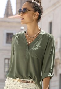 Olivegrüne Bluse mit langen Ärmeln, V-Ausschnitt, Knopfleiste und Brusttasche. Kombiniert mit hellen, gemusterten Hosen und übereinander getragenen Halsketten.