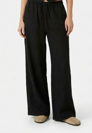 Žena oblečená v voľných čiernych nohaviciach s elastickým pásom a béžových plochých topánkach, stojaca na bielom pozadí.