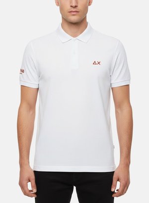 Uomo che indossa una polo bianca con piccoli loghi rossi sul petto sinistro e sulla manica destra, abbinata a pantaloni neri, su sfondo neutro.