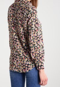 Blusa floreale con sfondo nero e motivi di margherite in rosa, giallo e bianco. Maniche lunghe con colletto standard, vestibilità non infilata.