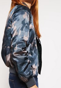 Osoba z długimi czerwonymi włosami, ubrana w granatowo-szary kamuflażowy kurtkę bombową i niebieskie dżinsy, stojąca bokiem na białym tle.