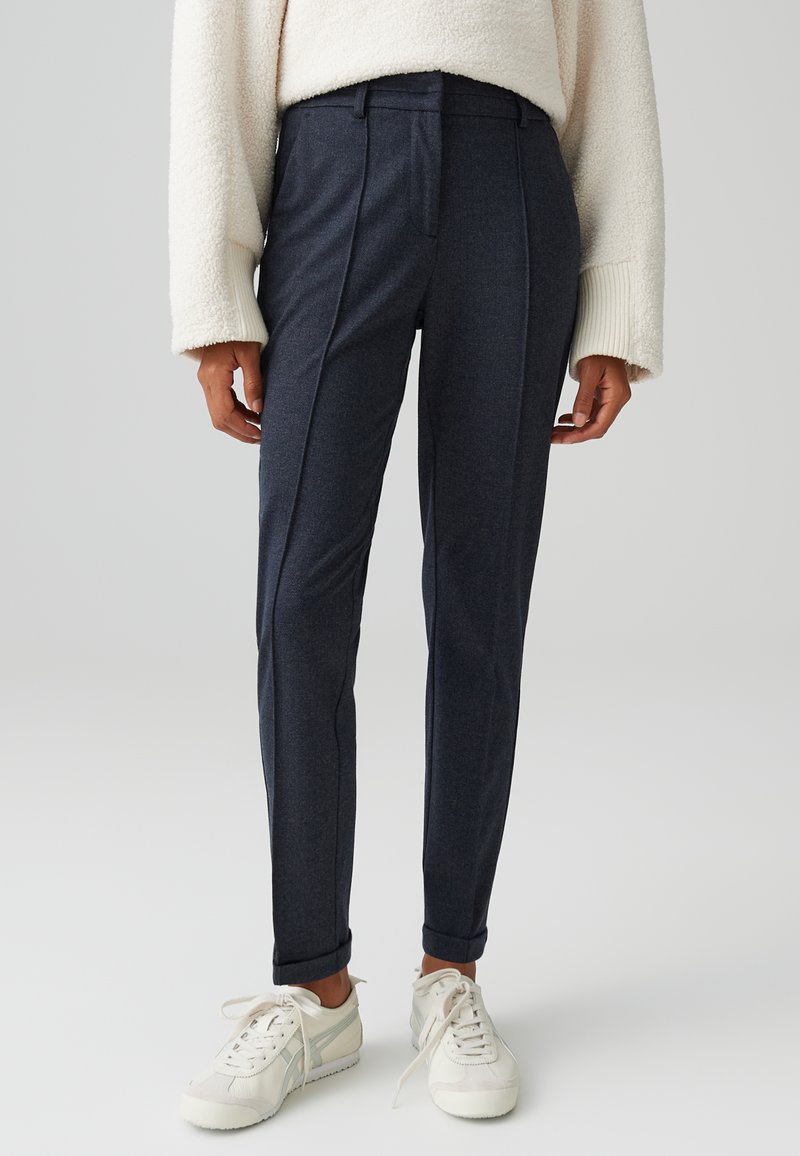 Pantaloni scuri blu su misura con una texture liscia, dotati di pieghe frontali, passanti per cintura e orli arrotolati. Abbinati a sneaker bianche.