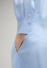 Hellblaues Kleid mit figurbetontem Design, kurzen Puffärmeln, Knöpfen an den Bündchen und seitlichen Taschen. Glatte Textur mit dezentem Glanz.