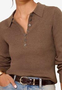 Chemise en maille côtelée marron avec un col à revers et une patte de boutonnage à cinq boutons. Associée à une ceinture foncée et un jean bleu clair.