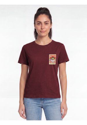 Femme portant un t-shirt bordeaux avec un motif de glace et un texte dans la zone de la poche poitrine, debout sur un fond blanc uni.