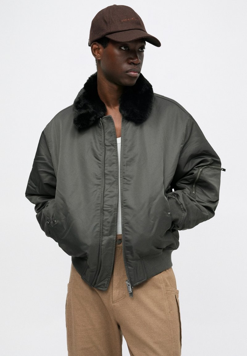Veste bomber verte avec un col en fausse fourrure, fermée par zip et dotée de poches latérales ; portée avec une casquette marron. Matière lisse, coupe décontractée.