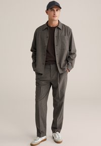 Lekka kurtka w kratę z brązowymi guzikami, zestawiona z pasującymi spodniami i ciemnym t-shirtem. Noszona z niebieską czapką i białymi trampkami.