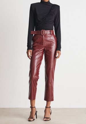 Nainen, jolla on musta pitkähihainen liehuva paita, korkeavyötäröiset viininpunaiset teksturoidut nahkahousut vyöllä ja mustat remmikkäät korkokengät.