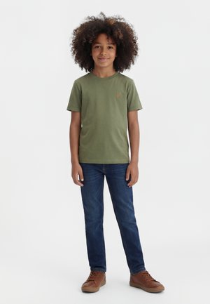 Enfant aux cheveux bouclés portant un t-shirt vert olive, un jean bleu et des baskets marron, debout devant un fond blanc uni.