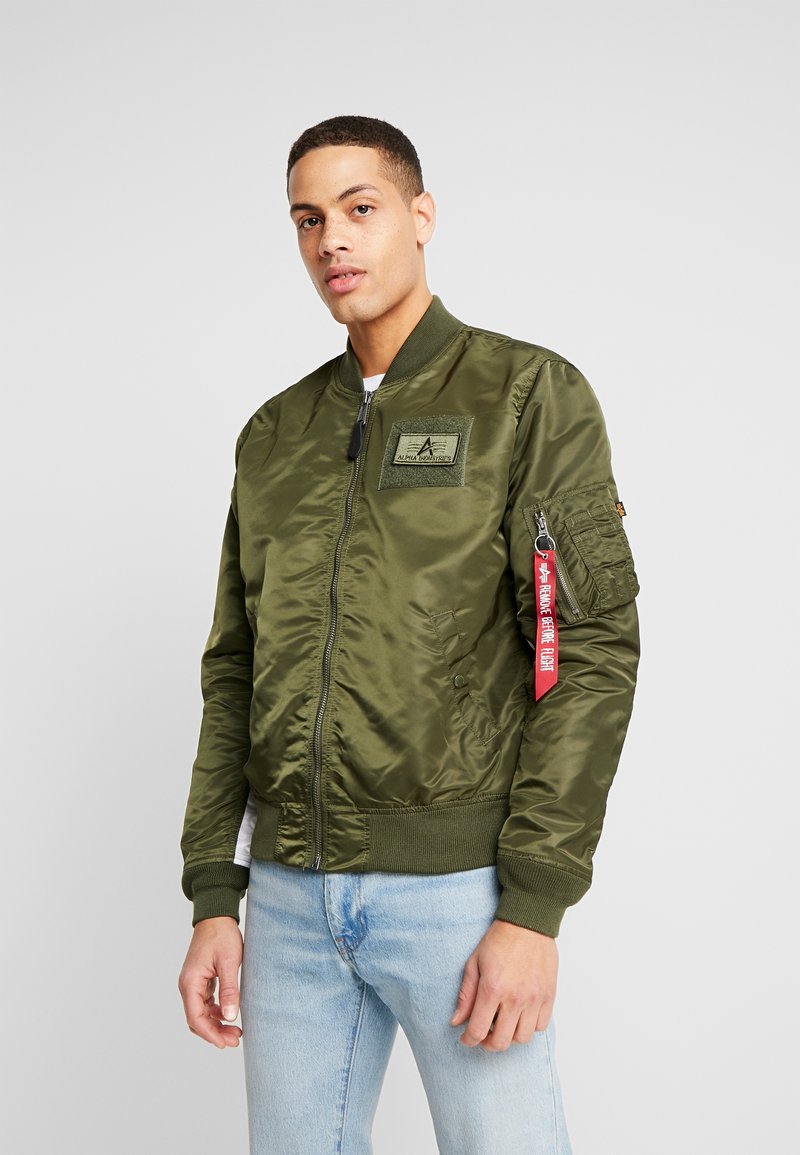 Grüne Satin-Bomberjacke mit Reißverschluss, gerippten Bündchen und einem strukturierten Patch auf der Brust; inklusive eines roten Tags am Ärmel.