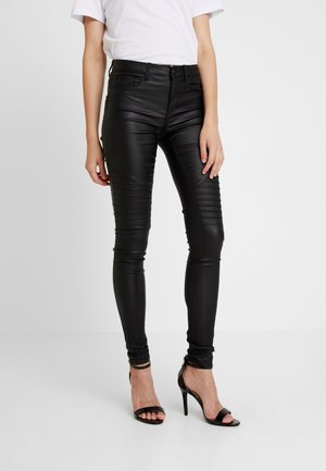 Femme portant un pantalon en cuir noir ajusté avec des détails côtelés aux genoux et des sandales à talons noirs, debout sur un fond blanc.