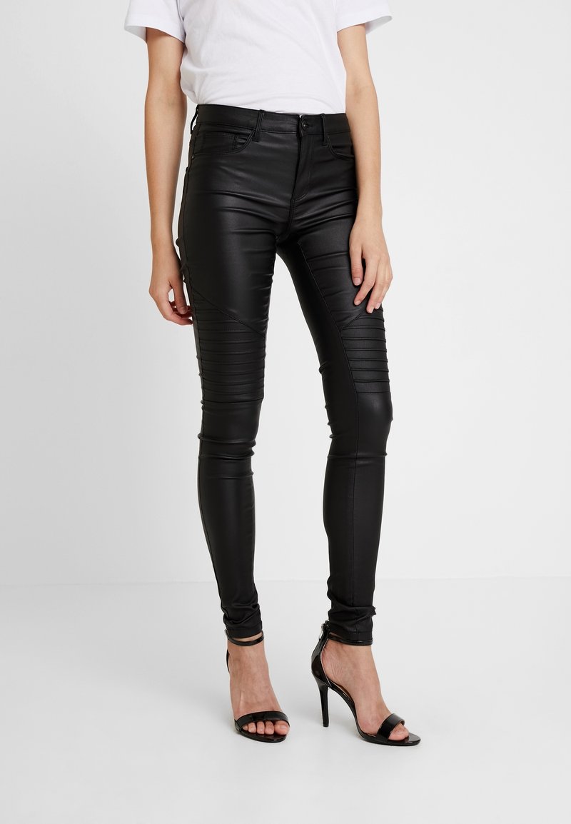 Femme portant un pantalon en cuir noir ajusté avec des détails côtelés aux genoux et des sandales à talons noirs, debout sur un fond blanc.