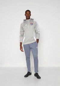 Pilkas sportinis džemperis su kapišonu ir baltais akcentais bei logotipu, derinamas su šviesiai pilkomis laisvalaikio kelnėmis. Abu drabužiai turi sportinį dizainą, pagaminti iš minkštos medžiagos.