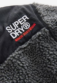 Black fabric panel with white "SUPERDRY®" text, red "SINCE 2003" and Japanese characters embroidered above textured dark gray fleece material.