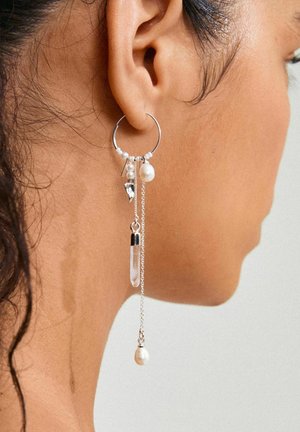Pendiente de aro plateado con múltiples elementos colgantes: perlas, un colgante de cristal y pequeñas cadenas. Presenta texturas mixtas y acentos brillantes.