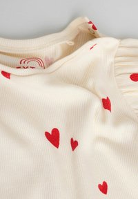 Krémový top z žebrované látky s červenými vzory srdce, s kulatým výstřihem a malým zapínáním na knoflíky vzadu.
