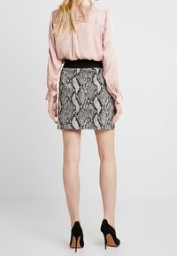 Blouse rose clair à manches longues associée à une mini-jupe noire à imprimé serpent et des escarpins noirs, mettant en valeur une silhouette près du corps et un tissu texturé.