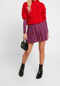 Czerwony sweter robiony na drutach z geometrycznym wzorem, fioletowa mini spódniczka w kwiaty, czarne botki za kostkę oraz mała czarna torebka.