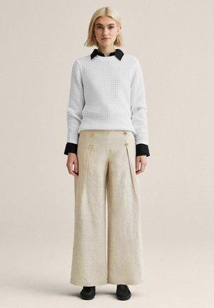 Lyshåret kvinde står iført en tekstureret hvid sweater over en sort skjorte med krave, beige bukser med vide ben og guldknapper, samt sorte sko.