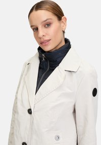 White trench coat with black buttons and peacoat collar, layered over a navy zip-up puffer. Features a black patch on the left sleeve.