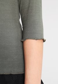 Olive green ribbed top with three-quarter sleeves; hem features a wavy edge detail. Material appears soft with a slight texture.